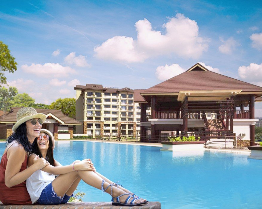 Two women enjoying a resort pool with a pavilion and multi-story building in the background.